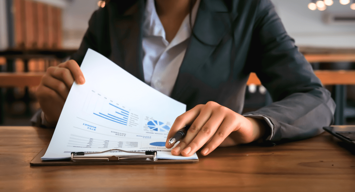 A person in a suite sits at a desk, visible from the neck down, as they look through a packet of paper as part of an audit process